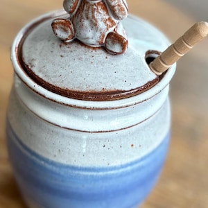 May include: A ceramic sugar bowl with a lid and a small wooden spoon. The bowl is white with a blue stripe and a brown rim. A small brown bear figurine sits atop the lid. The spoon is inserted into a hole in the lid.