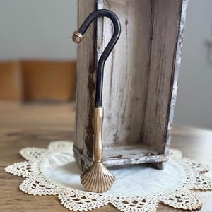 May include: A tall, rectangular, weathered white wooden mold with a hollow center stands upright. In front of it, a black metal hook with a gold-toned handle and a decorative gold-toned shell-shaped base rests on a white lace doily.