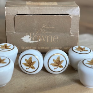 May include: Set of six white ceramic drawer knobs with a gold leaf design and gold trim. The knobs are from the "Towne" collection and are in their original cardboard box. The box reads "Hardware of Elegance".