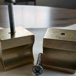 May include: Two brass-colored, square furniture feet with curved sides. One foot has a screw inserted, while the other has a hole. A loose screw is also present, suggesting a furniture repair or assembly project.