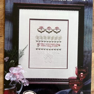 May include: Framed cross-stitch sampler with the text "Sumer is i-cumen in." The design features floral and geometric patterns in pink and green, with a teddy bear figure at the bottom. The frame is dark brown.