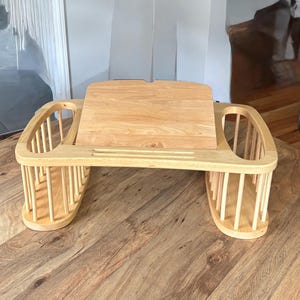 May include: A light-colored wooden lap desk featuring a central adjustable slanted surface for books or tablets, flanked by two integrated storage compartments with vertical spindle-style sides for holding items like pens, notebooks, or small accessories.