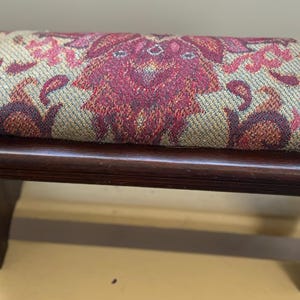 May include: A small, rectangular footstool with a dark brown wooden frame. The cushioned top features a tapestry design in shades of red, burgundy, and tan. The footstool has four legs and is suitable for use as a decorative accent or functional piece.