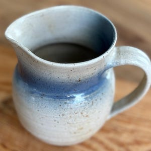 May include: A handmade ceramic pitcher with a rounded body and a curved handle. The pitcher features a gradient of colors, transitioning from a light beige at the base to a soft blue in the middle, and a white rim. The pitcher is ideal for serving beverages.