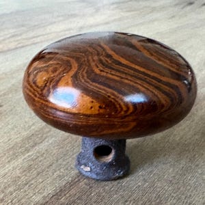May include: A round, polished cabinet knob featuring a glossy, swirling brown and dark wood grain pattern. The top is smooth and domed, while the base is a textured, dark gray metal cylinder with a visible circular opening for mounting hardware.