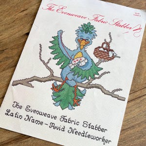 May include: A cross-stitch pattern featuring a blue bird with green wings and a yellow beak, perched on a brown branch. The bird is holding a needle and thread. The text "The Evenweave Fabric Stabber" and "Latin Name - Avid Needleworker" are printed below the image.