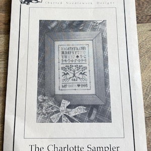 May include: A black and white printed needlework design titled "The Charlotte Sampler" by The Drawn Thread. The design features an alphabet, numbers, and a tree motif, framed within a wooden border. The text "her work 1995" is also included.