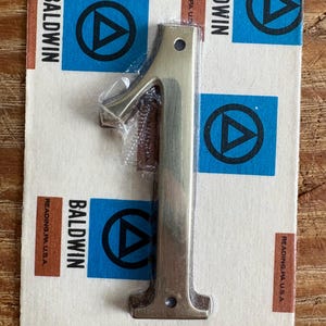 May include: A metallic number one door knocker, still in its original packaging. The packaging features the brand name "Baldwin" and a blue and black logo. The number is approximately 6 inches tall.