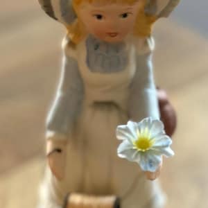 May include: A porcelain figurine of a young girl wearing a bonnet and holding a flower. The girl is dressed in a light blue dress and white apron. A spool of thread is attached to the figurine.