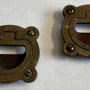 May include: Two antique brass drawer pulls with a D-shaped handle and four corner screws. The pulls have a weathered, aged appearance, suggesting they are vintage or reclaimed hardware. The hardware is designed for cabinets or drawers.