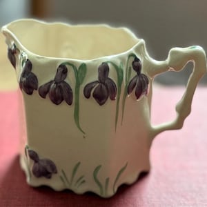 May include: Cream-colored ceramic creamer with a square shape and a curved handle. The creamer is decorated with purple flower and green stem designs. The flowers are painted on the front and sides of the creamer.