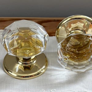 May include: Two door knobs featuring clear, multi-faceted glass handles shaped like large crystals. Each knob is mounted on a circular, polished gold-tone metal base plate, providing a reflective and metallic contrast to the transparent glass tops.