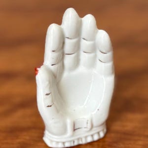 May include: A white ceramic hand sculpture with a detailed design. The sculpture is in an open palm pose, with visible finger and hand details. A small red accent is visible on the thumb. The sculpture is on a wooden surface.