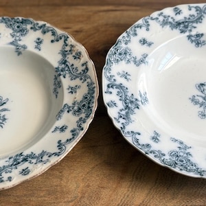 May include: Two white ceramic soup bowls with a blue floral pattern. The bowls have a scalloped edge and a central floral design. The blue pattern includes flowers and scrolling vines. The bowls are likely vintage or antique.