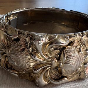 May include: Ornate silver-toned bracelet with detailed floral and scrollwork designs. The hinged bangle features a textured surface and a secure clasp. A vintage piece with a classic, elegant aesthetic.