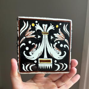 May include: A square ceramic tile featuring a black background with a central white symmetrical design of two stylized dolphins facing outward. The pattern includes white floral accents, small yellow dots, and a brown border frame.