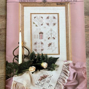May include: A framed needlework piece with a gold frame, featuring several cross-stitch designs. The text "CROSS 'N PATCH" is visible at the top. A white candle and a decorative cloth with fringe are in the foreground, along with roses and greenery.