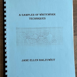 May include: A light blue spiral-bound book titled "A Sampler of Whitework Techniques" by Jane Ellen Balzuweit. The cover features an embroidered floral design. The book is on a wooden surface.