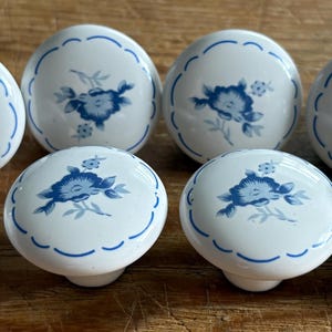 May include: Eight white ceramic cabinet knobs with a blue floral design. Each knob has a scalloped edge and a blue floral pattern on the top. The knobs are arranged on a wooden surface.