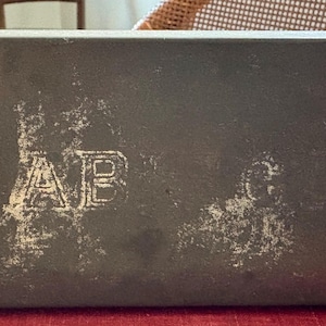 May include: A rectangular, dark gray metal box with a distressed finish. The box has the embossed letters "AB" on the front. The box is sitting on a red surface.