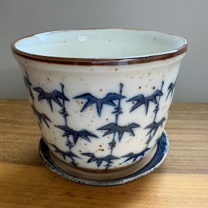 May include: A ceramic planter with a speckled off-white glaze and a repeating blue bamboo leaf pattern. The pot features a brown rim and sits on a matching circular saucer with a dark blue edge, showcasing a hand-crafted aesthetic.