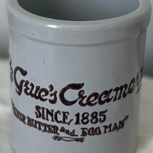 May include: A cylindrical, light gray stoneware crock with a dark brown inscription. The text reads "Grue's Creamery SINCE 1885 YOUR BUTTER and EGG MAN". The crock has a slightly speckled appearance and a smooth rim.