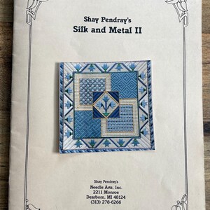 May include: A book cover titled "Shay Pendray's Silk and Metal II" featuring a detailed needlepoint design. The needlepoint is square, with shades of blue and white, and a central floral motif. The book is on a wooden surface.
