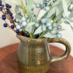 May include: A small, speckled, olive-green ceramic pitcher with a handle, filled with faux blue berries and light blue foliage. The pitcher is sitting on a wooden surface.