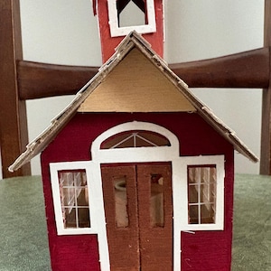 May include: A handcrafted miniature red schoolhouse with a white trim, a small steeple, and a wooden roof. The building features a double door, two windows, and a small porch. The schoolhouse is a charming decorative item.