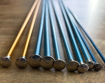 May include: A collection of knitting needles in various sizes. The needles have silver tips with size numbers, and the shafts are gold and blue. The needles are arranged on a wooden surface.