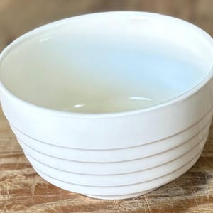 May include: A white ceramic bowl with a rounded shape and a series of horizontal, textured lines around the exterior. The bowl has a smooth, glossy finish and is set against a wooden surface.