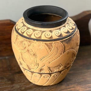 May include: A tan ceramic vase with a black rim and base. The body features a relief design of a winged lion in profile, surrounded by black scroll patterns at the top and bottom. The surface has a textured, matte finish with dark shading.