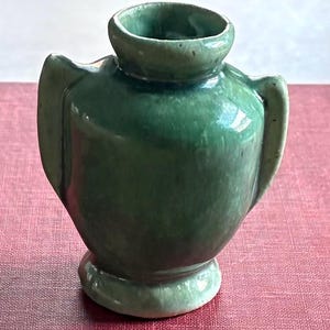 May include: A small, green ceramic vase with a glossy finish. The vase has a rounded body, a flared base, and two curved handles. The opening is circular and slightly raised. The vase is a deep, forest green color.