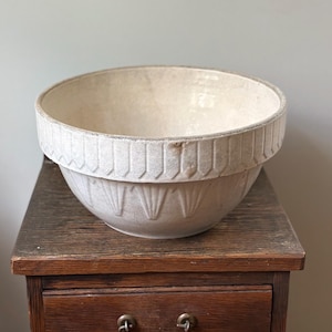 May include: A large, off-white ceramic mixing bowl featuring a wide, textured rim with vertical indentations and a lower body decorated with a repeating fan-like pattern. The bowl has a visible crackled glaze finish throughout its entire surface.