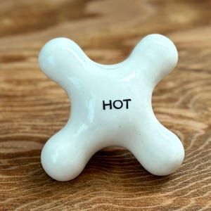 May include: A white, four-pronged ceramic faucet handle with a cross-shaped design. The center of the handle features the word HOT printed in clear, black, sans-serif capital letters. The surface of the handle is smooth, glossy, and rounded.