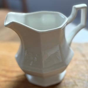 May include: A white ceramic creamer with a geometric design. The creamer has a handle and a spout for easy pouring. The creamer is sitting on a wooden surface. The creamer is a kitchenware item.