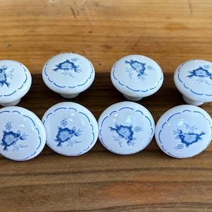 May include: Eight round white ceramic cabinet knobs arranged in two rows. Each knob features a blue floral pattern in the center and a thin blue scalloped border around the edge. The knobs have a smooth, glossy finish and a rounded dome shape.