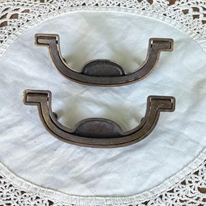 May include: Two matching semi-circular metal drawer pulls with a weathered bronze finish. Each handle features a curved, U-shaped design with a central raised tab and hinged ends that attach to a flat mounting surface, showing a dark patina.