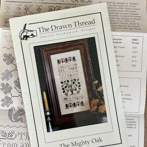 May include: A printed chart from "The Drawn Thread" for needlework designs, featuring a framed sampler with a tree and text. The chart includes the title "The Mighty Oak" and instructions. A candle holder and dried flowers are also visible.