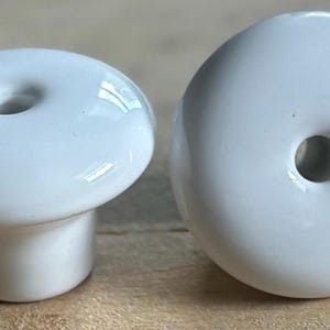 May include: Two white ceramic cabinet knobs. One knob is viewed from the side, showing a rounded top with a central hole and a cylindrical base. The other knob is viewed from the top, displaying a similar design. These knobs are suitable for furniture or home decor.