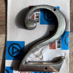 May include: A brushed metal number 2 door knocker. The number is mounted on a piece of paper with blue and white geometric shapes and the word "Baldwin". The knocker has two screw holes for mounting.