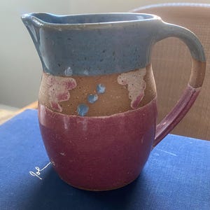 May include: A handmade ceramic pitcher with a blue, brown, and pink glaze. The pitcher has a wide mouth and a handle. It is decorated with a brown and pink design.
