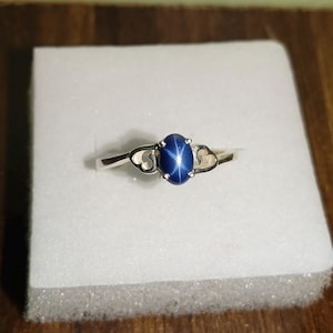 Könnte beinhalten: Ein silberner Ring mit einem großen, ovalen, dunkelblauen Edelstein. Der Ring liegt auf einer weißen Oberfläche, und das Band hat dekorative herzförmige Akzente auf beiden Seiten des Edelsteins. Der Ring wird auf einer Holzoberfläche präsentiert.