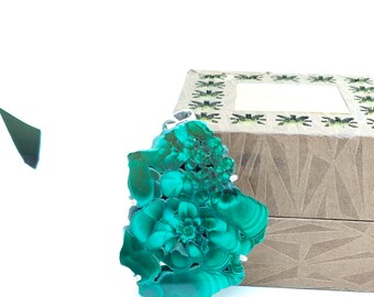 Pode incluir: Uma vibrante pedra de malaquite verde-azulado repousa ao lado de uma caixa decorativa. A pedra preciosa exibe padrões e texturas intrincados, mostrando sua beleza natural. A caixa é marrom clara com um design geométrico.