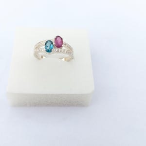 Könnte beinhalten: Ein silberner Ring mit zwei ovalen Edelsteinen, einem teal und einem magenta, nebeneinander angeordnet. Der Ring hat ein geteiltes Band mit kleinen, klaren Akzentsteinen. Der Ring wird auf einer weißen Oberfläche präsentiert.
