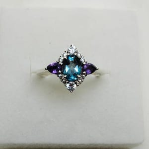 Könnte beinhalten: Ein silberner Ring mit einem ovalen, blauen Mittelstein, flankiert von zwei violetten Edelsteinen. Der Mittelstein ist von kleinen weißen Edelsteinen umgeben, die ein rautenförmiges Design bilden. Der Ring wird auf einer weißen Oberfläche präsentiert.