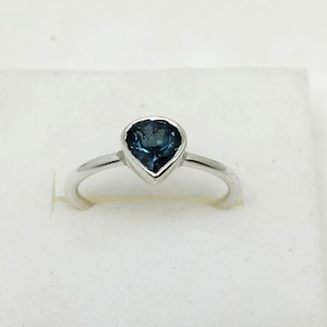 Könnte beinhalten: Ein silberner Ring mit einem tropfenförmigen, dunkelblau-grünen Edelstein. Der Ring hat ein poliertes Silberband, und der Edelstein ist in einer Lünettenfassung gefasst. Präsentiert auf einer weißen Oberfläche.