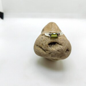 May include: A silver ring featuring a rectangular, light green gemstone. The ring is displayed on a small, textured, brown stone against a white backdrop. The gemstone is the central element of the ring's design.