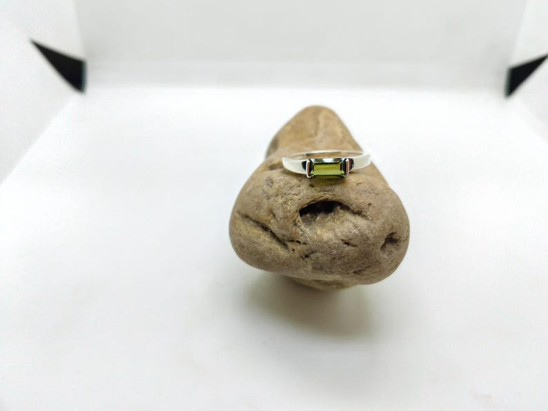 May include: A silver ring featuring a rectangular, light green gemstone. The ring is displayed on a small, textured, brown stone against a white backdrop. The gemstone is the central element of the ring's design.