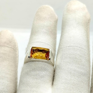 Könnte beinhalten: Ein silberner Ring mit einem großen, rechteckigen, goldgelben Edelstein. Der Ring wird vor einem weißen Hintergrund präsentiert, der das polierte Metall und die leuchtende Farbe des Steins hervorhebt. Die Facetten des Edelsteins fangen das Licht ein und erzeugen einen funkelnden Effekt.